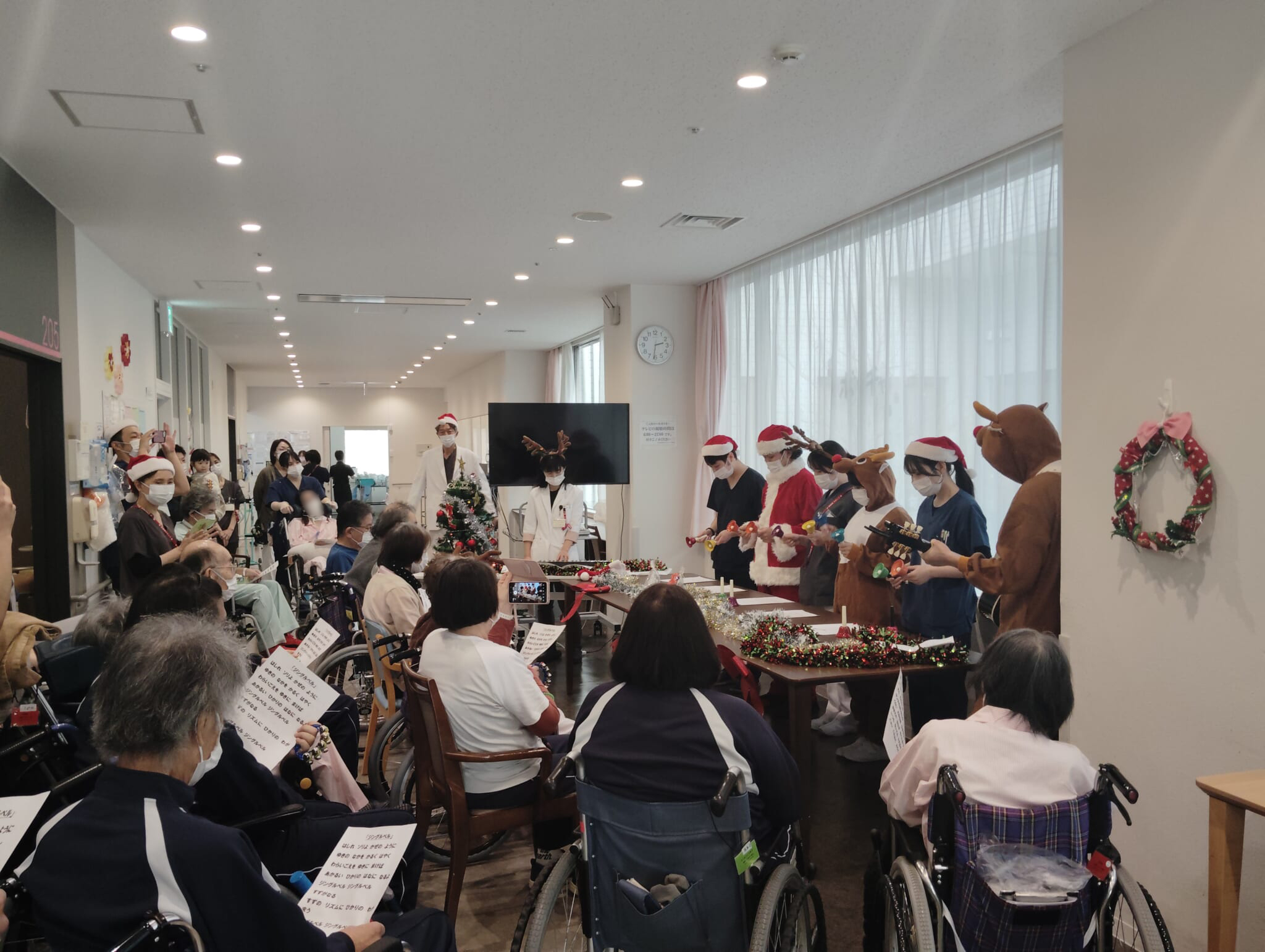 サンタクロースやトナカイに扮した職員がベルを演奏し、多くの患者さんが聞き入っている様子の写真