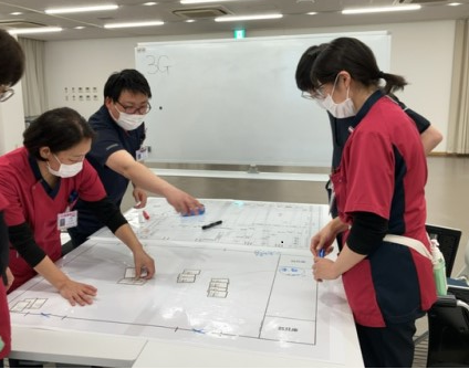 災害支援ナースと研修医が、相談しながら避難所運営ゲームで、大型の平面図に、避難者の「年齢・性別・国籍・持病・家族構成・ペットの有無」などが書かれたカードを配置している写真